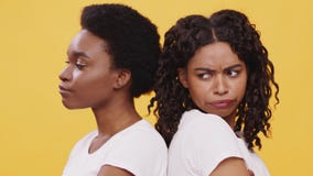 Two grumpy african american female friends standing back to back, feeling offended and irritated, orange background