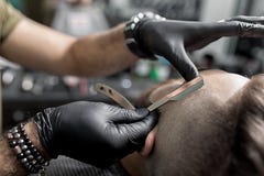Fryzjer męski w czarnych rękawiczkach żyłuje świątynię brutalny brodaty młody człowiek z prostą żyletką przy zakładem fryzjerskim zdjęcie royalty free Fryzjer męski w czarnych rękawiczkach żyłuje świątynię brutalny brodaty młody człowiek z prostą żyletką przy zakładem fryzjerskim zdjęcie royalty free