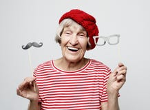 Funny grandmother with fake mustache and glasses, laughs and prepares for party over grey background stock photos Funny grandmother with fake mustache and glasses, laughs and prepares for party over grey background stock photos