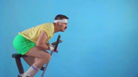 Funny young man from the 80`s with a mustache and glasses on exercise bike on a blue background slow mo stock footage Funny young man from the 80`s with a mustache and glasses on exercise bike on a blue background slow mo stock footage