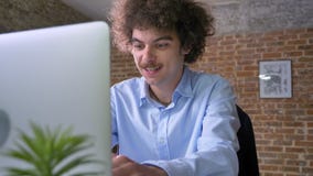 Funny young office worker with curly hair typing on laptop and sitting at table in modern office, smiling stock footage Funny young office worker with curly hair typing on laptop and sitting at table in modern office, smiling stock footage