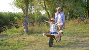 Férias em família alegres na natureza, o paizinho leva o menino alegre da criança em um carrinho de mão no movimento lento video estoque Férias em família alegres na natureza, o paizinho leva o menino alegre da criança em um carrinho de mão no movimento lento video estoque