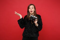Förvirrad ung kvinna i den fördelande handen för svart pälströja som rymmer den retro tappningfotokameran isolerad på ljust rött arkivbilder Förvirrad ung kvinna i den fördelande handen för svart pälströja som rymmer den retro tappningfotokameran isolerad på ljust rött arkivbilder