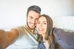 Gelukkig jong paar die een selfie met mobiele smartphonecamera nemen in de woonkamer die op bank thuis omhelzen royalty-vrije stock fotografie Gelukkig jong paar die een selfie met mobiele smartphonecamera nemen in de woonkamer die op bank thuis omhelzen royalty-vrije stock fotografie