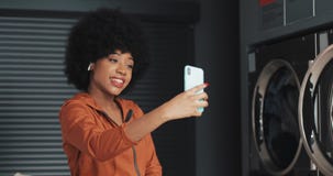 Gelukkige aantrekkelijke Afrikaanse Amerikaanse jonge vrouw die een videopraatje hebben bij laundromat Zelfbedienings openbare wa stock footage Gelukkige aantrekkelijke Afrikaanse Amerikaanse jonge vrouw die een videopraatje hebben bij laundromat Zelfbedienings openbare wa stock footage