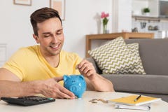 Gelukkige mens die muntstuk zetten in spaarvarken bij lijst in woonkamer stock foto Gelukkige mens die muntstuk zetten in spaarvarken bij lijst in woonkamer stock foto