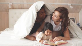 Genitori felici che leggono un libro al bambino allegro mentre coprendo di coperta stock footage Genitori felici che leggono un libro al bambino allegro mentre coprendo di coperta stock footage