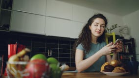 Giovane bella donna abbastanza caucasica riccia che si siede alla tavola in cucina piacevole facendo uso dello smartphone, sta ma archivi video Giovane bella donna abbastanza caucasica riccia che si siede alla tavola in cucina piacevole facendo uso dello smartphone, sta ma archivi video