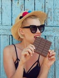 Giovane donna attraente in occhiali da sole ed in cappello di paglia che mangia cioccolato su un fondo blu Efect del film fotografia stock Giovane donna attraente in occhiali da sole ed in cappello di paglia che mangia cioccolato su un fondo blu Efect del film fotografia stock