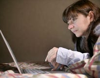 Giovane donna castana sveglia che si trova sul letto e che scrive in suo computer portatile immagine stock Giovane donna castana sveglia che si trova sul letto e che scrive in suo computer portatile immagine stock