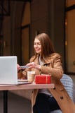 Giovane donna che si siede in una caffetteria Prende il suo dessert sul telefono Sulla tavola è il computer portatile all'esterno fotografie stock Giovane donna che si siede in una caffetteria Prende il suo dessert sul telefono Sulla tavola è il computer portatile all'esterno fotografie stock