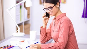 Giovane stilista femminile attraente che lavora alla scrivania, dissipante mentre comunicando sul mobile fotografia stock libera da diritti Giovane stilista femminile attraente che lavora alla scrivania, dissipante mentre comunicando sul mobile fotografia stock libera da diritti