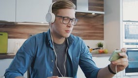 Giovane uomo di risata che guarda scorrendo film sullo smartphone che mangia i cereali con le cuffie Giovane uomo di risata che guarda scorrendo film sullo smartphone che mangia i cereali con le cuffie