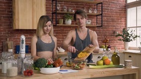 Giovani coppie in vestiti di sport che stanno dietro il tavolo da cucina che prepara sull'insalata fresca del lunc con le verdure archivi video Giovani coppie in vestiti di sport che stanno dietro il tavolo da cucina che prepara sull'insalata fresca del lunc con le verdure archivi video