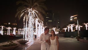 Girl and boy walking on the street the night Dubai. Among the glowing palms trees. UAE.