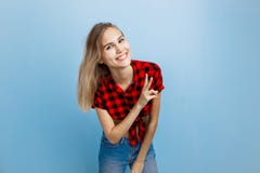 Gladlynt stilig ung blond flicka med blåa ögon som bär den röda rutiga skjortan och jeans som visar tecken V, anseende royaltyfri foto Gladlynt stilig ung blond flicka med blåa ögon som bär den röda rutiga skjortan och jeans som visar tecken V, anseende royaltyfri foto