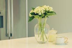 Glass vas av vita blommor och tekoppen i ett kök med suddigt royaltyfri bild Glass vas av vita blommor och tekoppen i ett kök med suddigt royaltyfri bild