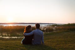Glückliche Paare, die auf dem Gras aufpasst den Sonnenuntergang vor einem See sitzen lizenzfreie stockfotografie Glückliche Paare, die auf dem Gras aufpasst den Sonnenuntergang vor einem See sitzen lizenzfreie stockfotografie
