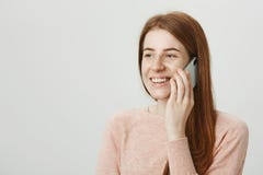 Good-looking redhead female talking on smartphone while looking aside and smiling cheerfully, standing over gray stock photography Good-looking redhead female talking on smartphone while looking aside and smiling cheerfully, standing over gray stock photography