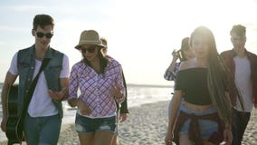 Group of young hipster friends walking together on a beach at the water`s edge holding a guitar. Slowmotion shot stock video Group of young hipster friends walking together on a beach at the water`s edge holding a guitar. Slowmotion shot stock video