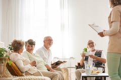 Gruppo di pensionati senior di casa di cura che si siedono insieme al salone comune che ascolta il giovane infermiere fotografia stock libera da diritti Gruppo di pensionati senior di casa di cura che si siedono insieme al salone comune che ascolta il giovane infermiere fotografia stock libera da diritti