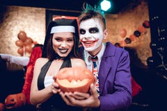 Halloween party. A guy in a Joker costume and a girl in a nun costume posing with a pumpkin-lamp. Behind them sit their friends and relax Halloween party. A guy in a Joker costume and a girl in a nun costume posing with a pumpkin-lamp. Behind them sit their friends and relax