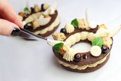 Hand med pincett som avslutar lantliga cirklar för Paris Brest chouxbakelse med hasselnötganache, muttrar, äppleskivor som är sva royaltyfri fotografi Hand med pincett som avslutar lantliga cirklar för Paris Brest chouxbakelse med hasselnötganache, muttrar, äppleskivor som är sva royaltyfri fotografi