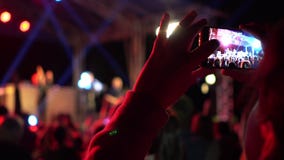 Hands of girl making video of summer festival concert with mobile phone. Crowd celebrating party event. Defocused image