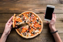 Hands of a woman ordering pizza with a device over a wooden workspace table. All screen graphics are made up.