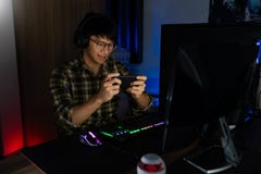 Handsome Excited Asian Gamer Guy In Headphones Enjoy And Rejoicing While Playing Video Games On Computer In Cozy Room Is Lit With Royalty Free Stock Photos