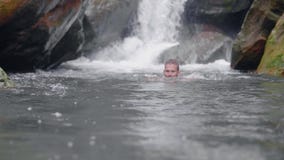 Handsome man enjoying swim in mountain river from tropical waterfall in rainforest. Young man bathing in rocky river on stock footage Handsome man enjoying swim in mountain river from tropical waterfall in rainforest. Young man bathing in rocky river on stock footage