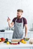 handsome smiling bearded man in apron listening music in earphones and holding chili pepper while cooking