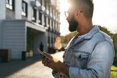 Handsome smiling hipster businessman with beard, in denim jacket, trendy glasses walks around city and holding smartphone stock photos Handsome smiling hipster businessman with beard, in denim jacket, trendy glasses walks around city and holding smartphone stock photos