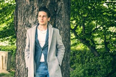 Handsome young man leaning against tree. Looking at camera, in a sunny day wearing a light grey coat Royalty Free Stock Photos Handsome young man leaning against tree. Looking at camera, in a sunny day wearing a light grey coat Royalty Free Stock Photos
