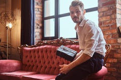 A handsome young man with a stylish beard and hair elegantly dressed holding a gift box while sitting on a red vintage stock photography A handsome young man with a stylish beard and hair elegantly dressed holding a gift box while sitting on a red vintage stock photography