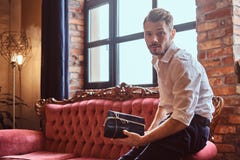 A handsome young man with a stylish beard and hair elegantly dressed holding a gift box while sitting on a red vintage stock image A handsome young man with a stylish beard and hair elegantly dressed holding a gift box while sitting on a red vintage stock image