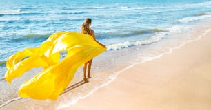 Happy Carefree Woman walking on Sea Beach with flying Long Yellow Fabric as Sail. Beautiful Girl enjoying Sun Tan on Summer
