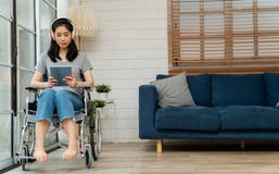 Happy Disabled Asian Woman Sitting In A Wheelchair And Working With Tablet At Home, The Concept Of Technologies For The Stock Photos
