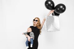 Happy elegant young mom, girl in black dress happily holding bags with black balloons, and little baby on white background. Baby