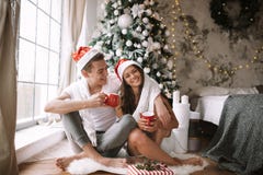 Happy guy and girl in white t-shirts and Santa Claus hats sit with red cups on the floor in front of the window next to royalty free stock photos Happy guy and girl in white t-shirts and Santa Claus hats sit with red cups on the floor in front of the window next to royalty free stock photos