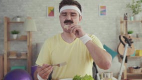 Happy and joyful young sporty man with a mustache eating vegetables sitting at the table stock video footage Happy and joyful young sporty man with a mustache eating vegetables sitting at the table stock video footage
