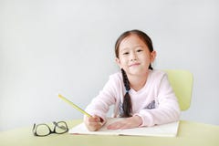 Happy little Asian child girl write in a book or notebook with pencil sitting on kid chair and table against white background