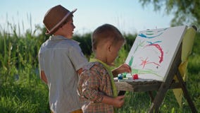 happy male child together with his younger brother draw with brushes and colored paints on canvas while resting outdoors