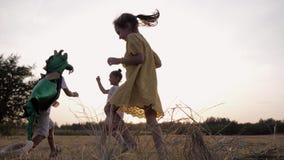 Happy pastime, male child in dragon costume plays an active game with female friends running around meadow during sunset
