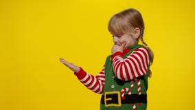Kid girl in Christmas elf Santa helper costume points fingers at blank space shows advertising area