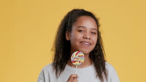 Studio portrait of positive african teen girl licking rainbow lollipop, enjoying sweets and smiling, orange background