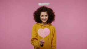 Happy Valentine`s Day! Angelic girl afro hairstyle with nimbus holding pink heart, looking playful flirting and sending air kiss,