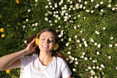 Happy woman listening to audiobook while lying on grass among blooming daisies, top view