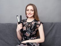 Happy young adorable woman in long dress on grey sofa holding hair styler up at home against grey wall stock photos Happy young adorable woman in long dress on grey sofa holding hair styler up at home against grey wall stock photos