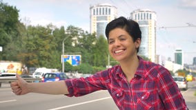 Happy young woman with black short hair lifting thumb and looking for taxi, standing on street near road in urban city stock video Happy young woman with black short hair lifting thumb and looking for taxi, standing on street near road in urban city stock video